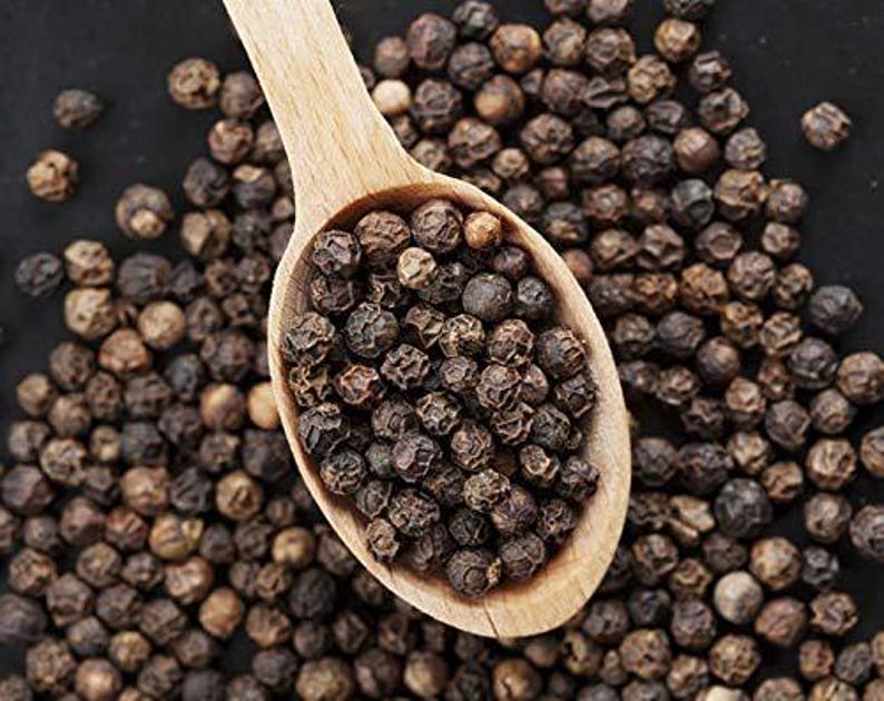 May include: A wooden spoon filled with whole black peppercorns, with more peppercorns scattered around it. The peppercorns are dark brown and round, and the spoon is light brown.