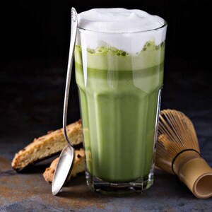 May include: A tall glass of matcha latte with a thick layer of white foam, a silver spoon, and biscotti. A bamboo whisk rests beside the glass. The drink is a vibrant green color, and the background is dark.