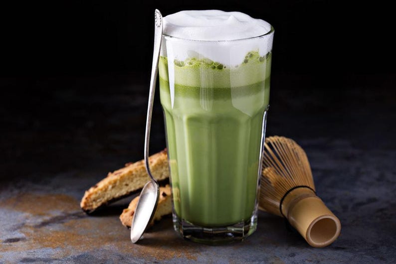May include: A tall glass of matcha latte with a thick layer of white foam, a silver spoon, and biscotti. A bamboo whisk rests beside the glass. The drink is a vibrant green color, and the background is dark.