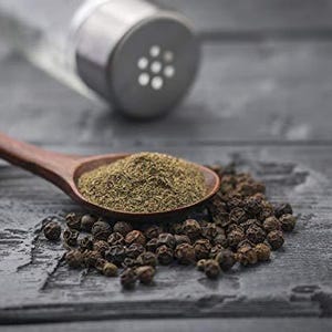 May include: A wooden spoon filled with ground black pepper sits next to a pile of whole peppercorns. A clear glass pepper shaker with a metal top is in the background. The scene is set on a dark, textured surface.