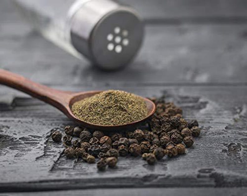 May include: A wooden spoon filled with ground black pepper sits next to a pile of whole peppercorns. A clear glass pepper shaker with a metal top is in the background. The scene is set on a dark, textured surface.