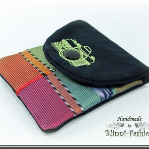 Pode incluir: Uma pequena bolsa de tecido preto com um forro de tecido listrado colorido. A bolsa tem uma aba com um design de câmera bordado em verde e um fecho de botão preto. O texto "Handmade by Blinni-Fashion" está impresso abaixo da bolsa.