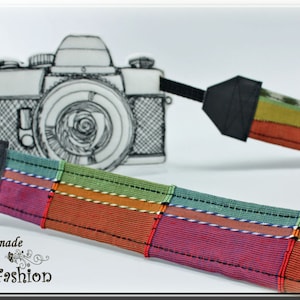 Pode incluir: Uma correia de câmera colorida com um detalhe em couro preto e uma imagem desenhada à mão de uma câmera vintage. A correia é feita de diferentes tecidos coloridos com um padrão listrado. O texto "Handmade by Blinni-Fashion" está na parte inferior da imagem.