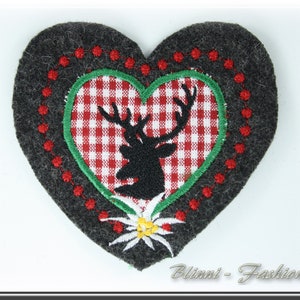 Op de afbeelding: Een grijs vilten hartvormig patch met een rood en wit geruit hart erin. Het geruite hart heeft een groene rand en een zwart silhouet van een hert met geweien. De buitenrand van de patch is een rij rode stippen.