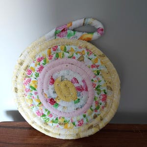 May include: A round, yellow, pink, and white quilted hot pad with a floral pattern. The hot pad has a loop handle.