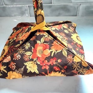 May include: A fabric casserole carrier featuring a dark brown background with a vibrant floral print in shades of orange, yellow, and red. The carrier has four triangular flaps that fold upward and are secured by a mustard yellow handle strap.