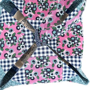 May include: A quilted pillow with a patchwork design. The pillow features a pink floral pattern, navy blue and white gingham, and teal leaf patterns. The pillow has a pinwheel design with four sections that meet in the center.