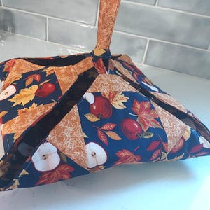 May include: A fabric bowl cozy featuring a dark blue background with a pattern of red apples, apple slices, and autumn leaves in shades of orange and yellow. The square-shaped holder has four folded corners and a single upright fabric handle.