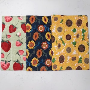 May include: Three fabric placemats with different patterns. The first placemat is green with red strawberries, the second is navy blue with yellow sunflowers, and the third is white with yellow sunflowers.