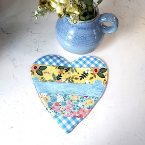 May include: A blue and white patchwork heart-shaped coaster with a floral pattern. The coaster is made of fabric and has a quilted texture.