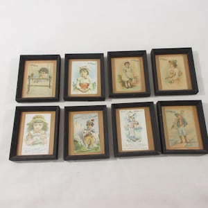 May include: A collection of eight rectangular black frames, each containing a vintage-style trade card featuring color illustrations of children in various outfits and settings, with some cards displaying printed text and brand names.