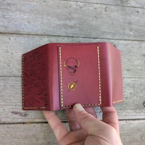 May include: A maroon leather tri-fold wallet featuring yellow contrast stitching. The center panel displays a black outline of a superhero mask above a yellow lightning bolt emblem. The left panel is embossed with a repeating pattern of lightning bolts.