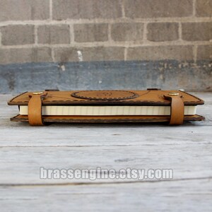 May include: A brown leather journal with a decorative design and a white paper insert. The journal has a stitched edge and a leather strap closure.