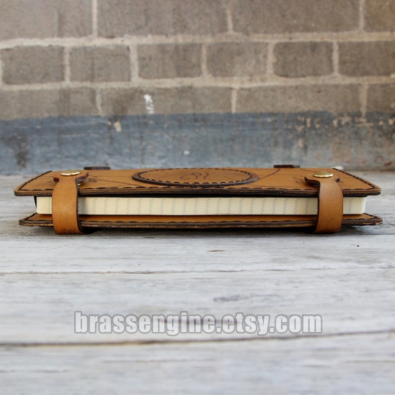 May include: A brown leather journal with a decorative design and a white paper insert. The journal has a stitched edge and a leather strap closure.