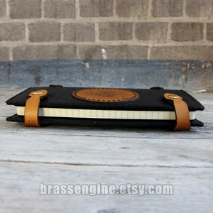 May include: A black leather journal with a brown leather strap and a circular design on the front. The journal is closed and lying on a wooden surface.