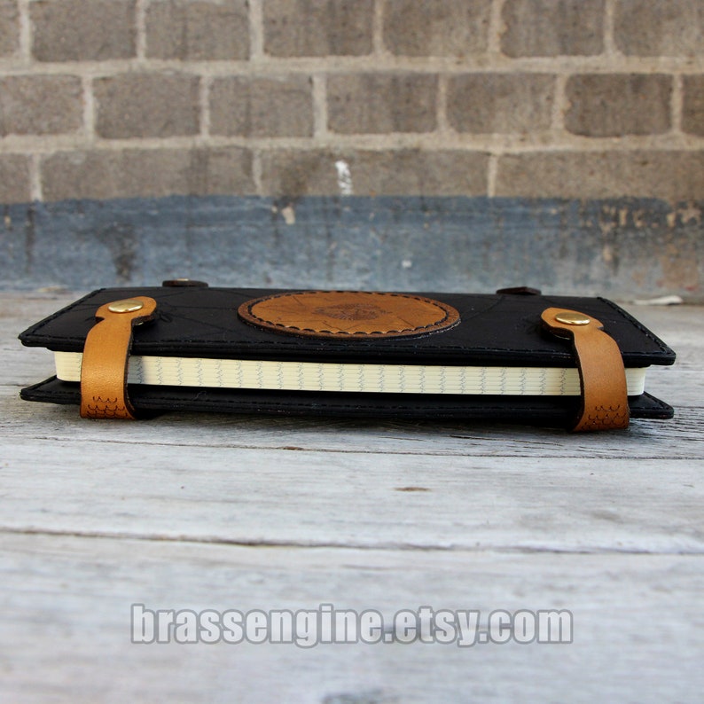 May include: A black leather journal with a brown leather strap and a circular design on the front. The journal is closed and lying on a wooden surface.