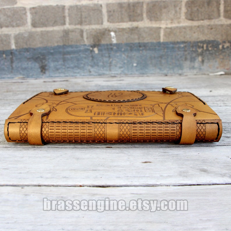 May include: A brown leather journal with an intricate design and a brass clasp. The cover features an embossed pattern of hieroglyphs and a circular design with a stylized bird.