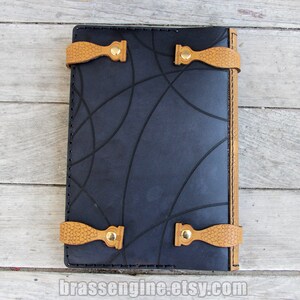 May include: Black leather journal with a geometric pattern embossed on the cover. The journal has tan leather straps with brass buckles and a stitched edge.