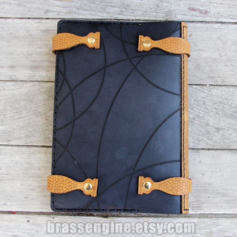 May include: Black leather journal with a geometric pattern embossed on the cover. The journal has tan leather straps with brass buckles and a stitched edge.