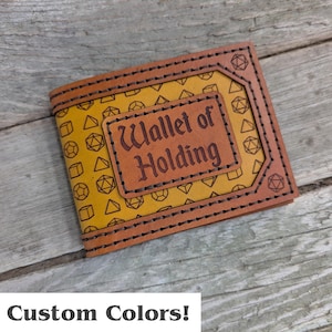 May include: A brown leather wallet with the text "Wallet of Holding" embossed on a rectangular patch. The wallet features a yellow interior with geometric shapes and is stitched with dark thread. The phrase "Custom Colors!" is at the bottom.