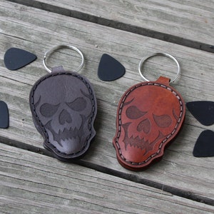 May include: Two leather keychains with a skull design. One keychain is brown and the other is a reddish brown. Both keychains have a silver key ring.