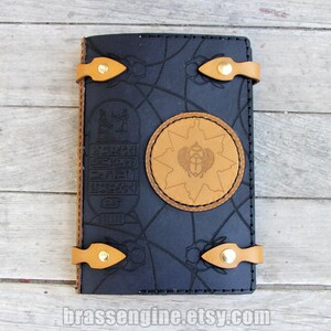 May include: Black leather journal with a brown leather starburst design featuring an Egyptian scarab beetle. The journal has two brown leather straps with brass buckles.