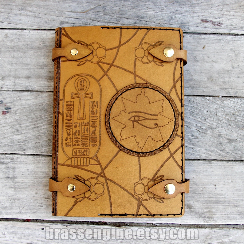 May include: A brown leather journal with an intricate design featuring an Egyptian ankh symbol, the Eye of Horus, and a starburst. The journal has two leather straps with brass buckles.