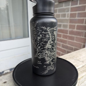 May include: A black insulated water bottle with a map design. The bottle has a black lid with a flip-top opening. The map is etched in a lighter color, showing geographical features. The bottle is sitting on a black round coaster.
