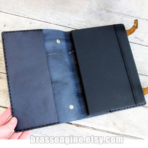 May include: A black leather notebook cover with a flap and a pen loop. The cover is open to reveal a black notebook with a blank page.