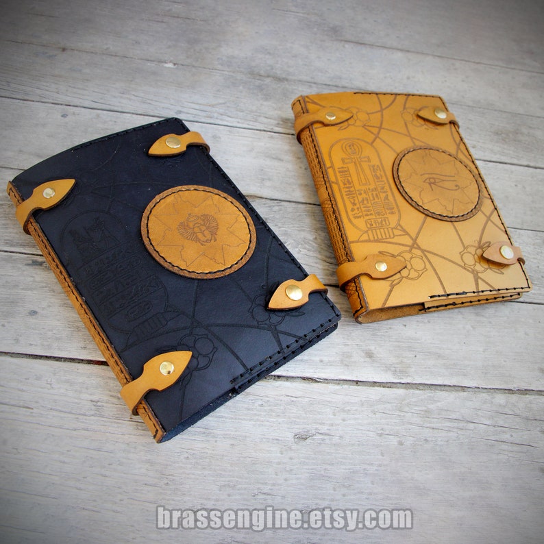 May include: Two leather-bound journals with intricate designs. One is black with gold accents and the other is brown with gold accents. Both feature Egyptian-inspired motifs, including hieroglyphs and an ankh symbol.