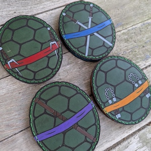 May include: Four green coasters with a turtle shell design. Each coaster features a different colored band and weapon design: red with sais, blue with swords, purple with a staff, and orange with nunchucks. The coasters are on a wooden surface.