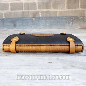 May include: A black leather journal with a gold embossed design and brown leather straps. The journal is closed and lying on a wooden surface.