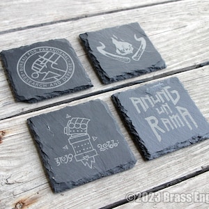 May include: Four black slate coasters with different designs. The first coaster has a circle with the text "BUREAU FOR PARANORMAL RESEARCH AND DEFENSE". The second coaster has a flame design. The third coaster has a hand with a symbol and the text "2988:". The fourth coaster has the text "ANUNG UN RAMA".