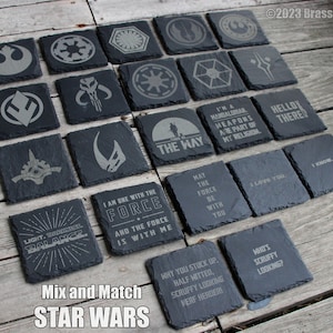 May include: A set of 16 black slate coasters with various Star Wars themed designs, including logos, characters, and quotes. The coasters are arranged in a grid pattern on a wooden surface.