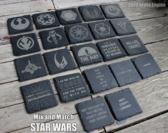 May include: A set of 16 black slate coasters with various Star Wars themed designs, including logos, characters, and quotes. The coasters are arranged in a grid pattern on a wooden surface.
