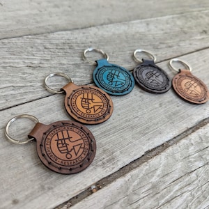 May include: Five leather keychains with a brown, blue, and black color scheme. Each keychain features a circular design with the text "Bureau for Paranormal Research and Defense" and a symbol.