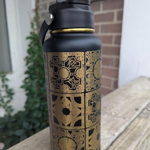 May include: A matte black insulated water bottle featuring a gold-colored geometric puzzle box pattern printed on the exterior. The bottle has a black plastic lid with a carry handle and a gold-colored metal ring accent located just below the bottle cap.