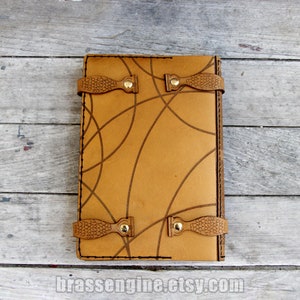 May include: A brown leather journal with a geometric pattern and two leather straps with brass buckles. The journal is closed and lying flat on a wooden surface.
