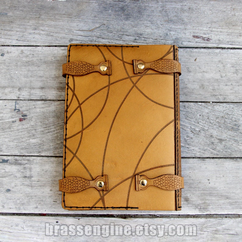 May include: A brown leather journal with a geometric pattern and two leather straps with brass buckles. The journal is closed and lying flat on a wooden surface.