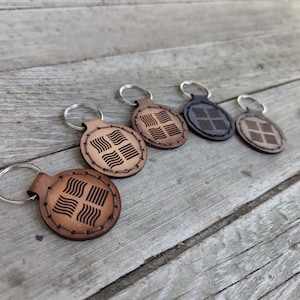 May include: Five leather keychains with a repeating wave pattern design. The keychains are brown, dark brown, and black. The keychains are on a wooden surface.