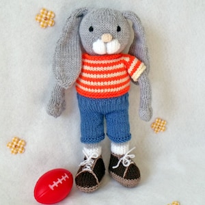 May include: A gray knitted bunny rabbit wearing a striped orange and white shirt, blue jeans, and brown shoes. The bunny is holding a red football.