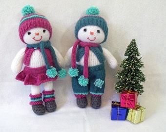 May include: Two knitted snowman dolls wearing pink and teal winter outfits. The dolls are standing in front of a small artificial Christmas tree with red, blue, and yellow wrapped gifts.
