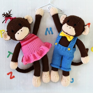 Puede incluir: Dos juguetes de mono de punto colgando de una percha blanca. El mono de la izquierda lleva un vestido rosa con un dobladillo con volantes. El mono de la derecha lleva un mono azul con una pajarita amarilla.