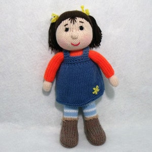 May include: A knitted doll with brown hair, wearing a blue dress with a yellow flower, an orange sweater, and brown shoes.