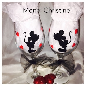 May include: Two clear wine glasses decorated with black silhouettes of a mouse couple and red heart accents. Each glass has a black bow at the base and is filled with white tissue paper. The text "Mone' Christine" is above the glasses.