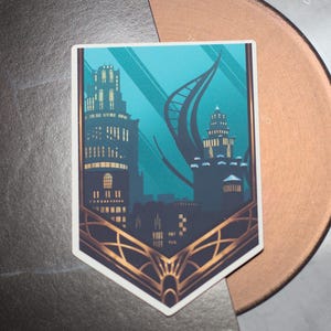 Puede incluir: Una pegatina con una ilustración estilizada de un horizonte de ciudad con dos edificios, uno alto y delgado, el otro más bajo y ancho. Los edificios están delineados en oro y colocados sobre un fondo azul verdoso. La pegatina está sobre una superficie negra.