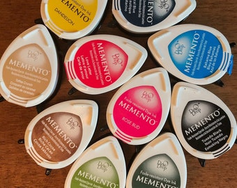 May include: A collection of ten Memento Ink Pads in various colors, including Toffee Crunch, Rose Bud, Nautical Blue, Bahama Blue, Tuxedo Black, Rich Cocoa, Bamboo Leaves, Northern Pine, and Dandelion. Each ink pad features the Memento logo and the ink color name.