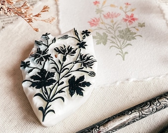 May include: A white, carved, floral stamp with black ink. The stamp features a design of stylized flowers and leaves.