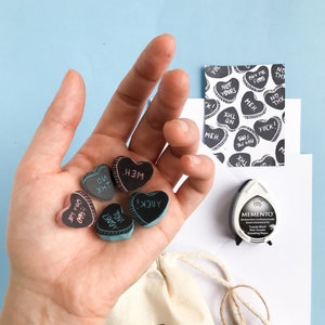 May include: A set of six heart-shaped rubber stamps with various phrases, including "YOU XHT", "MEH", "I  JUY", "BUY ME FORD", and "YUCK!" The stamps are in a hand, and there is a small bag with the stamps printed on it. The bag has a brown tag that says "HAND MADE" in a circle.