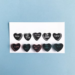 May include: A set of 10 black heart-shaped candy conversation hearts with various messages written in white, pink, and blue ink. The messages include "MEH", "NO THX", "BUY ME FOOD", "NOT YOURS", "YUCK!" and their reversed versions.